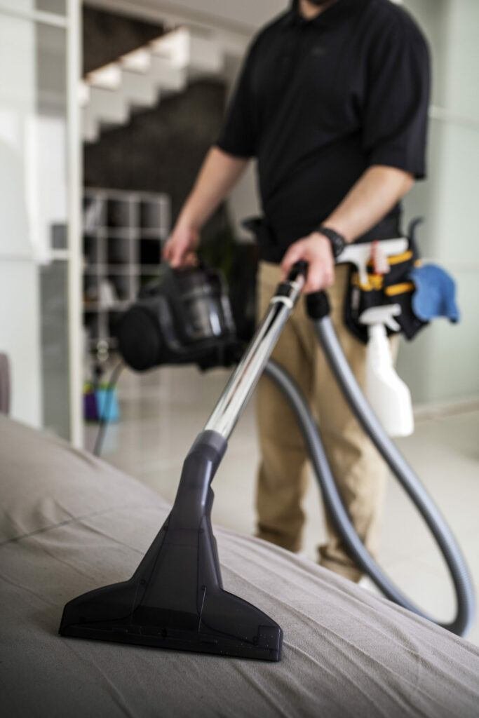 A man using a vacuum cleaner to clean a bed at Commerce City